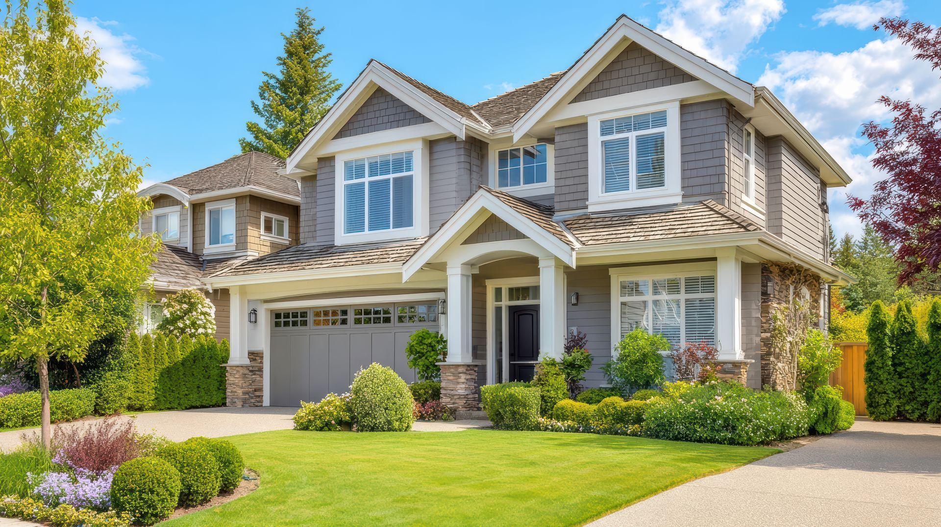 Suburban two-story house with beige siding, white trim, manicured lawn, and a driveway under a blue sky