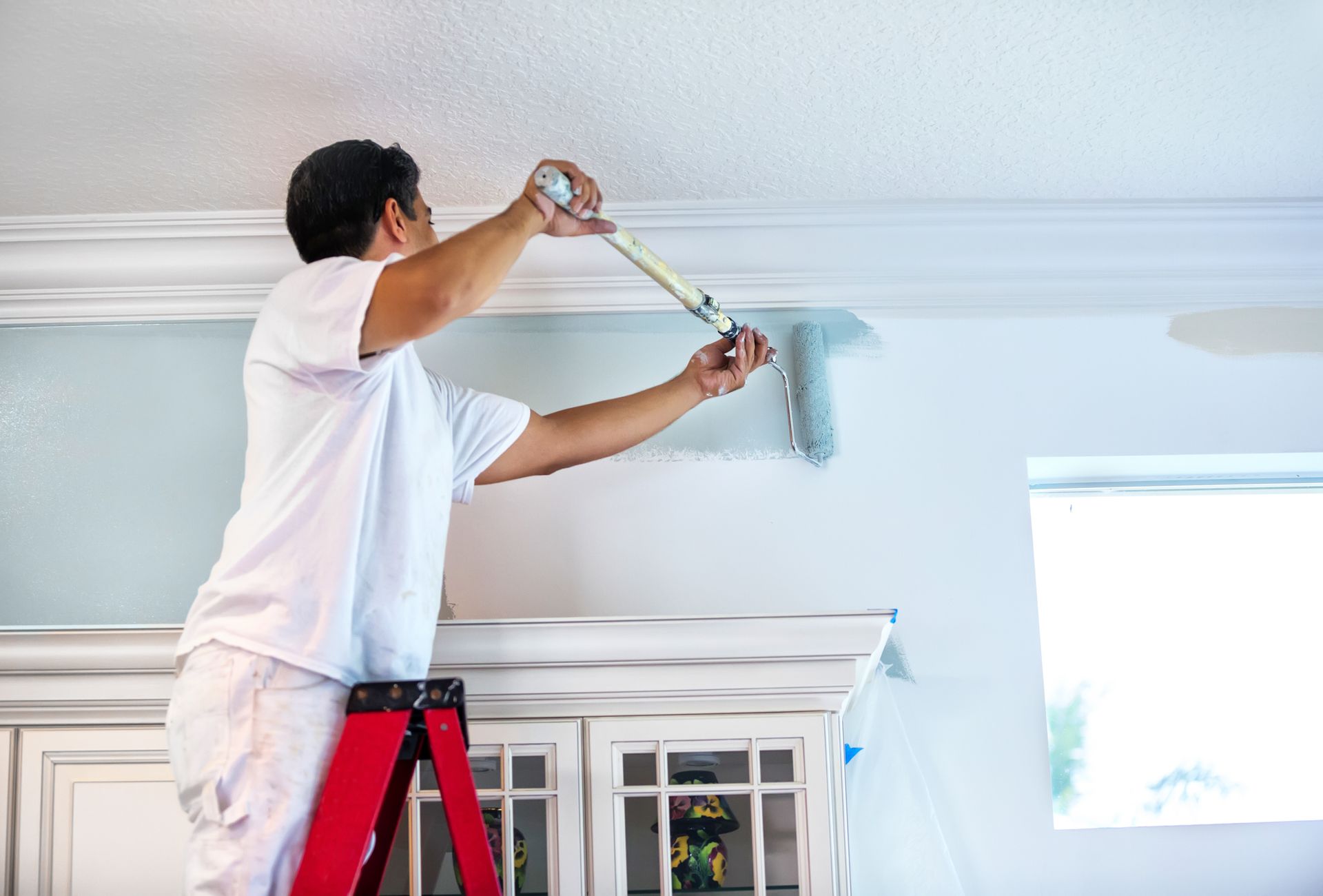 Man painting a white ceiling with a roller while standing on a red ladder in a bright room