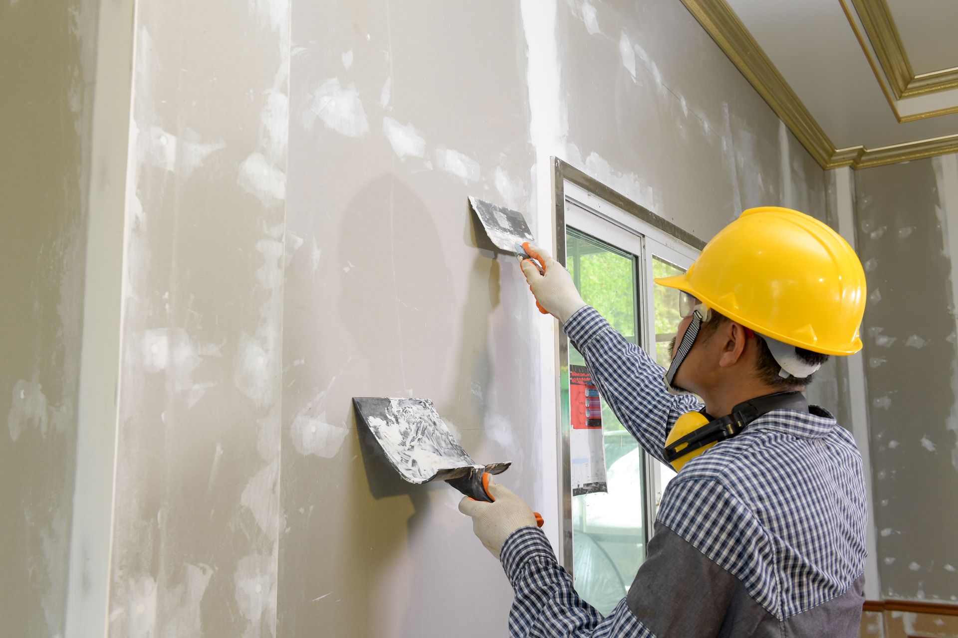 Worker in yellow hard hat smoothing drywall with a trowel inside a room under renovation.