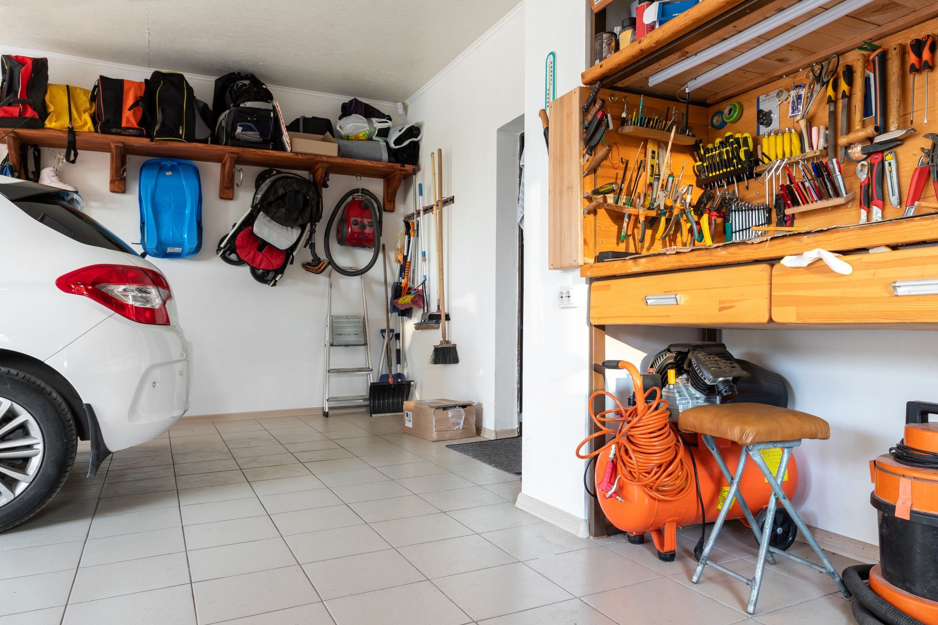 Garage interior with a white car, wall-mounted tools, shelves, and a wooden workbench with orange gear