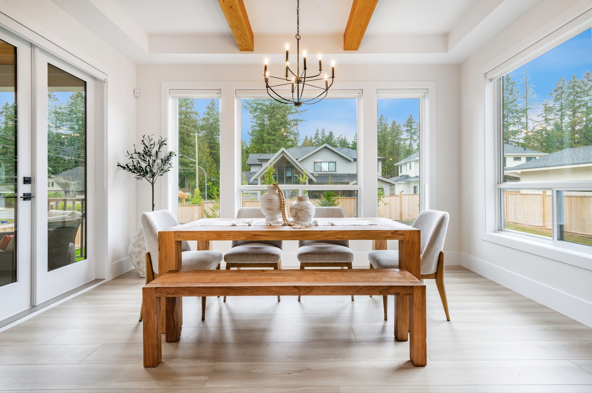 Bright dining room with wooden table, bench, white walls, and large windows overlooking a yard