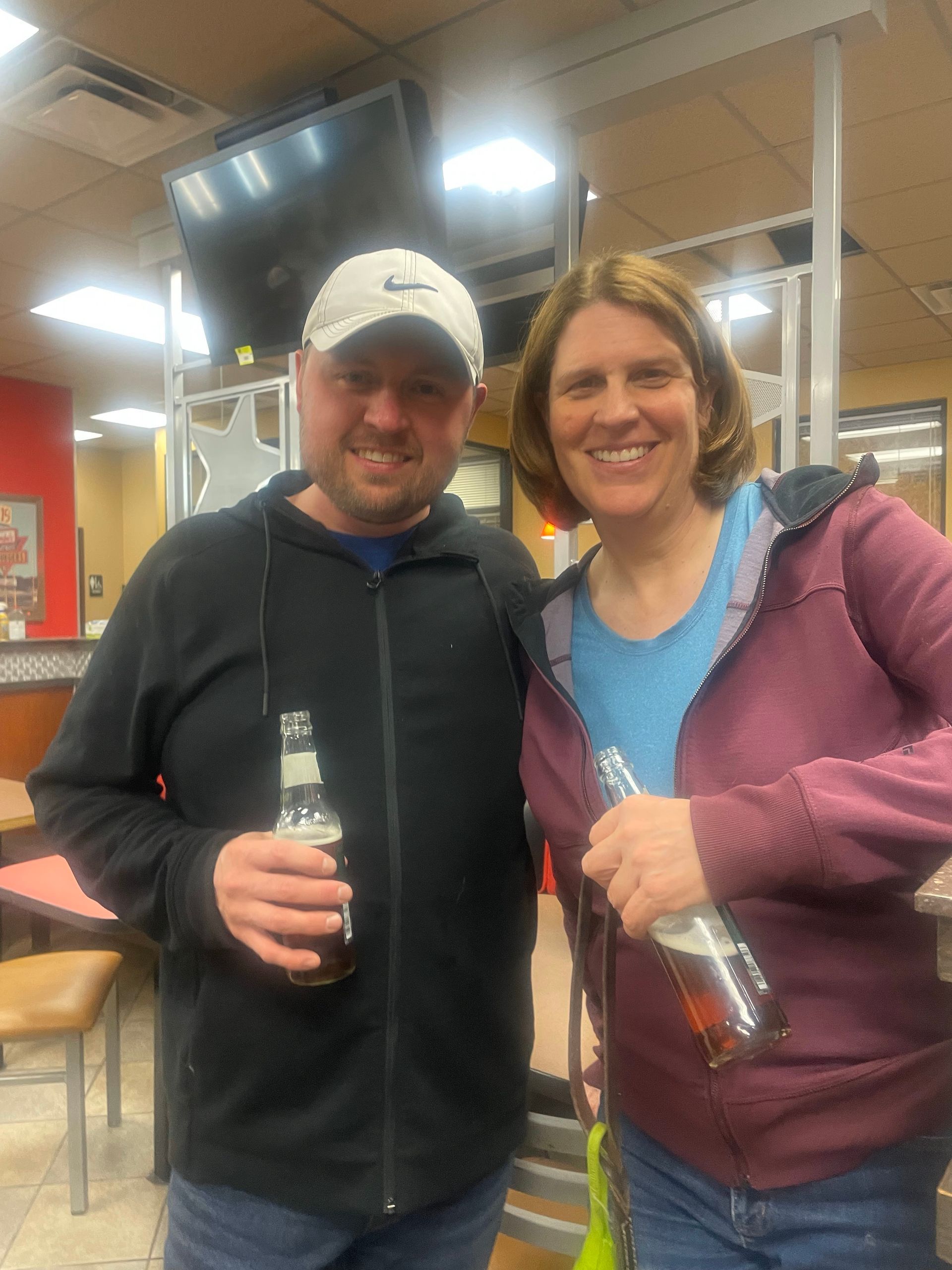 Two people smiling, holding bottles, indoors. Man in black jacket and cap, woman in purple jacket and blue shirt.