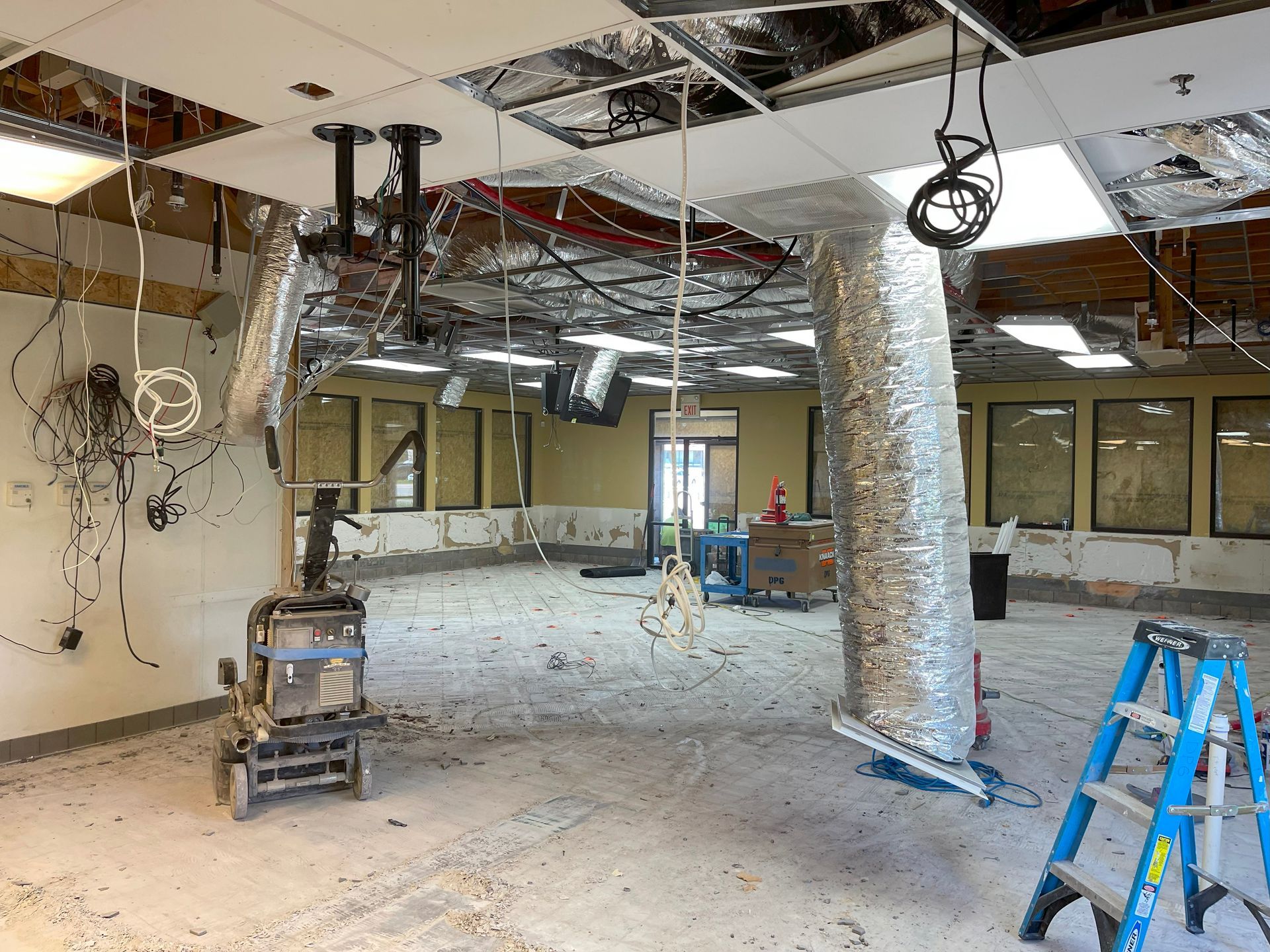 Interior room under renovation; exposed ceiling, ductwork, wires. Blue ladder, debris on floor, and brick walls.