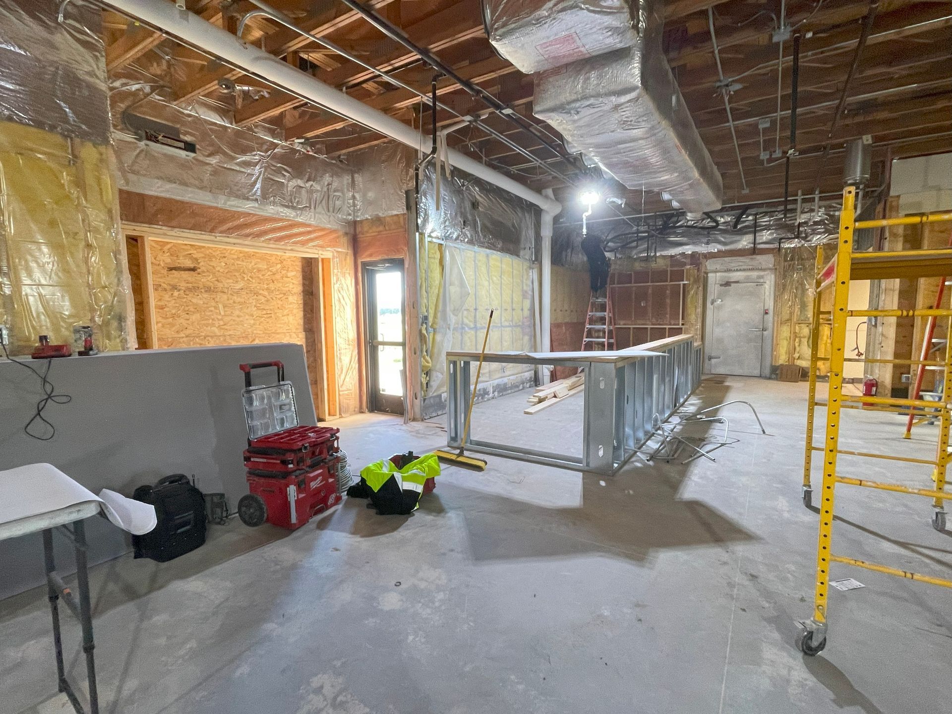 Interior construction site with exposed ceiling, framing, and unfinished walls. Scaffolding, tools, and a partial bar are visible.