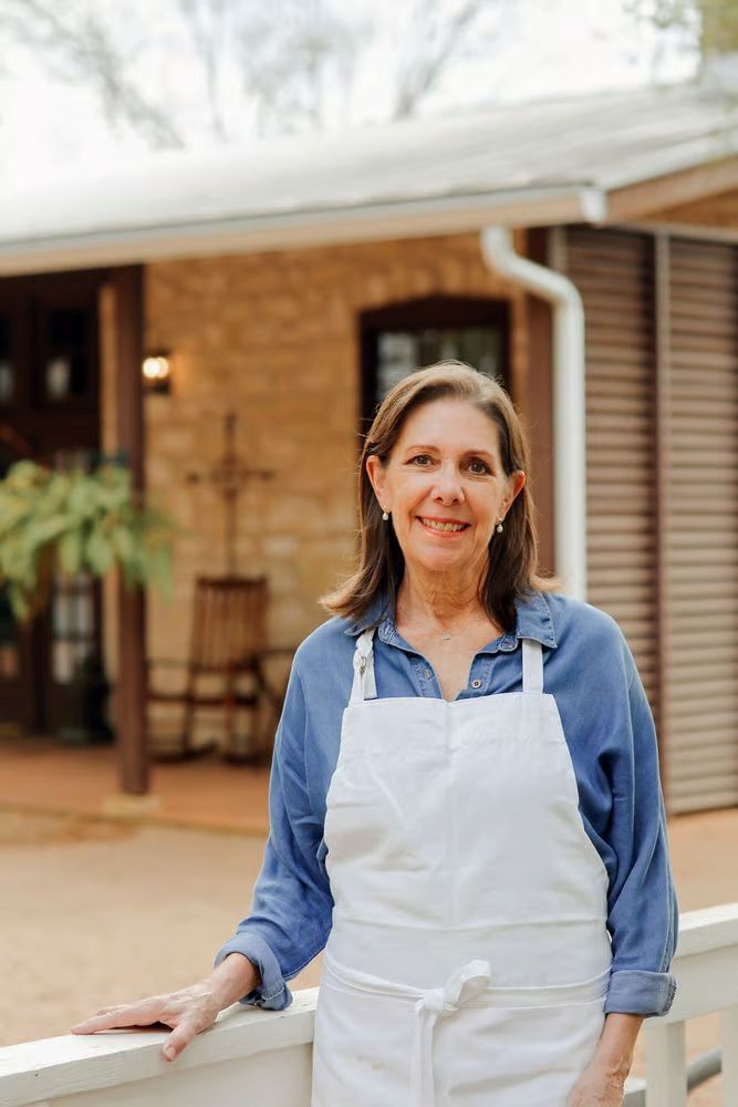 A woman in an apron is leaning on a white fence in front of a house.