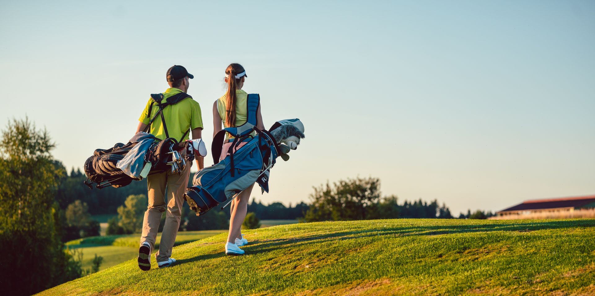 Two golfers with bags walking across a grassy hill under a bright sky.