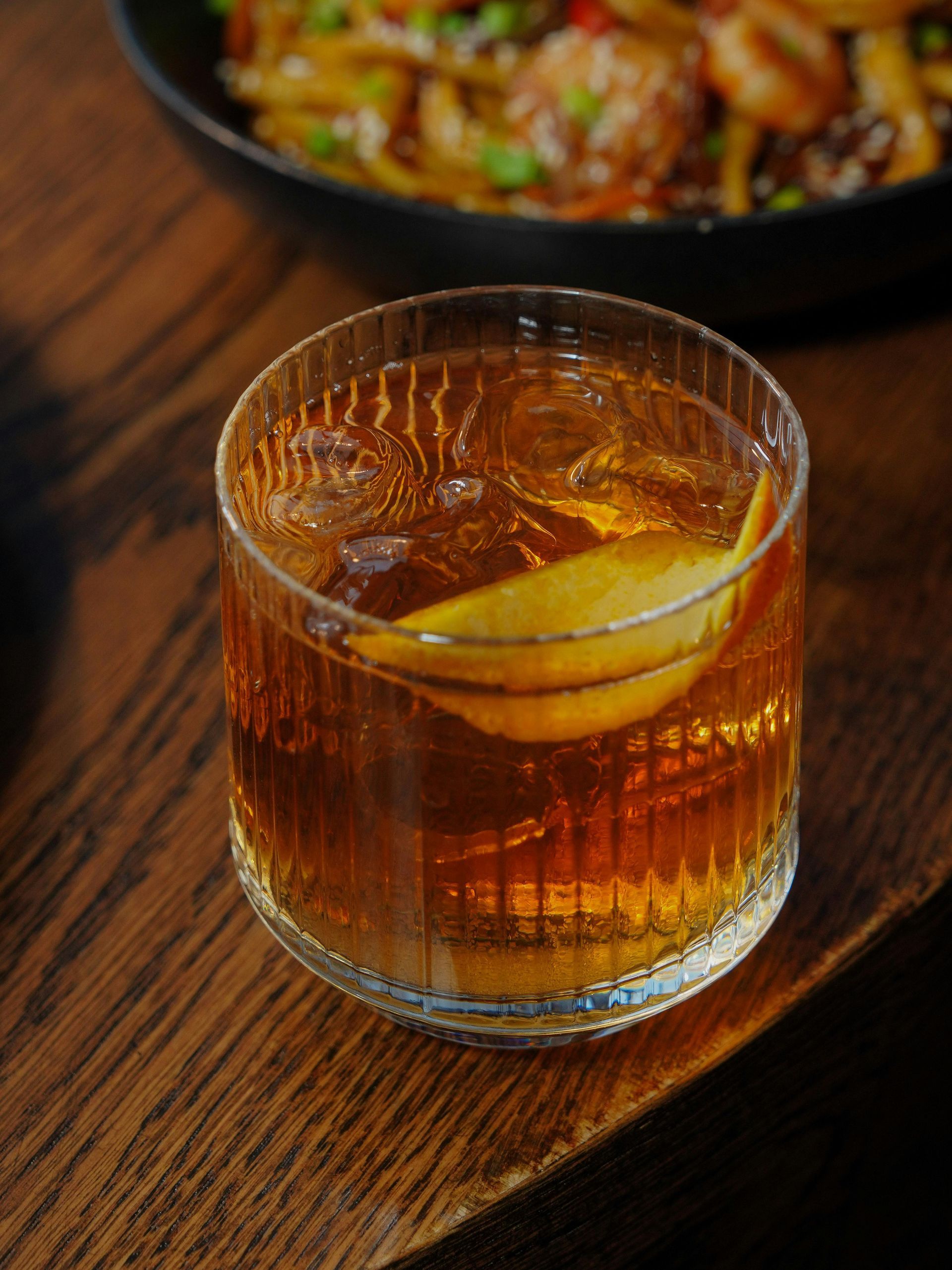 A glass of whiskey with ice and a lemon peel on a wooden table.