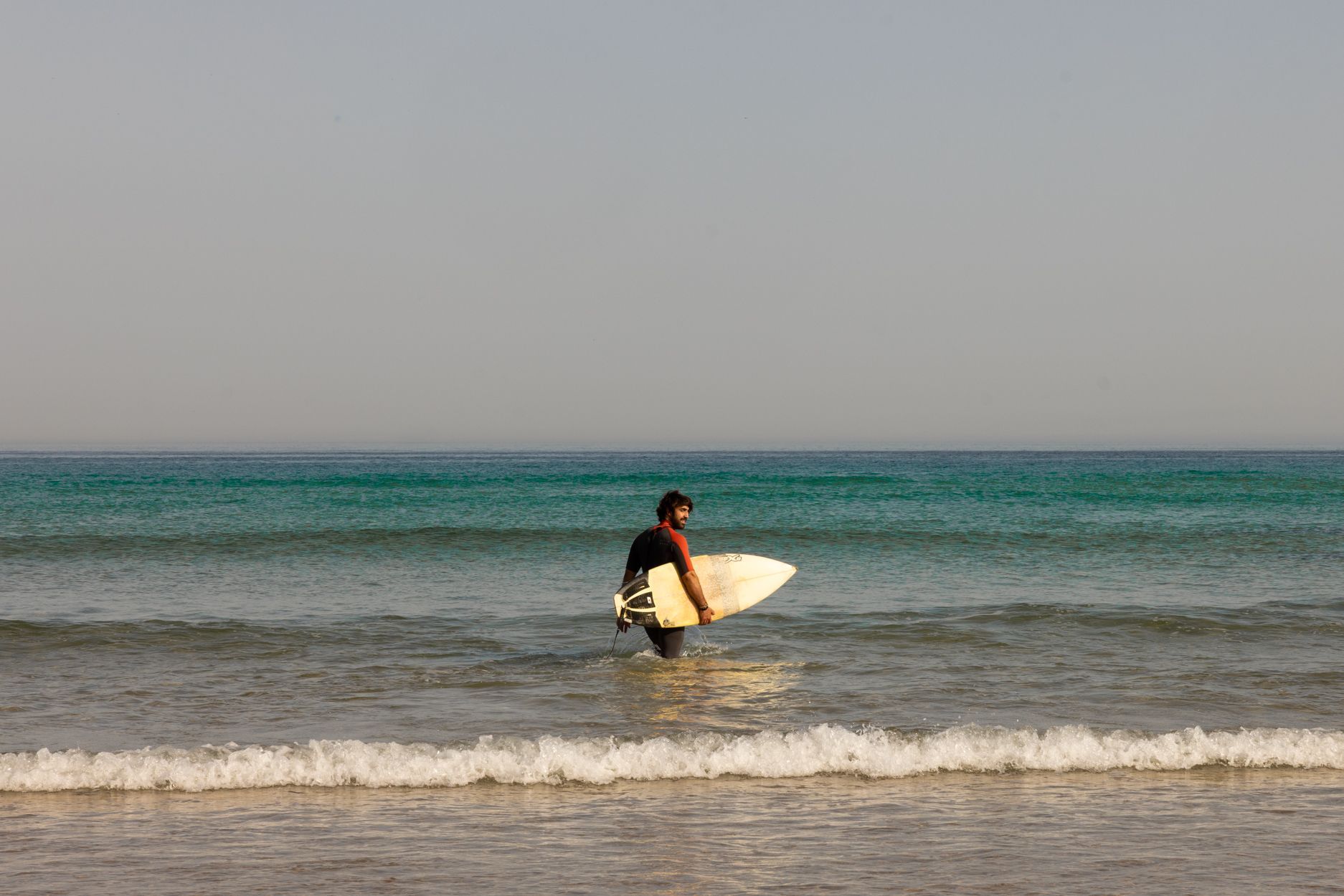 Un surfista lleva una tabla de surf en aguas poco profundas del océano, con olas y un horizonte tranquilo.