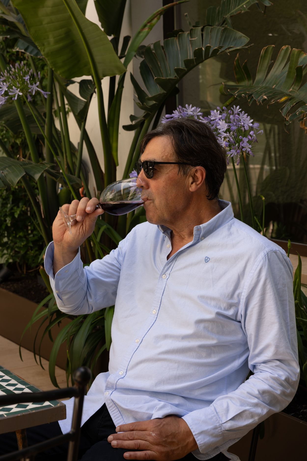 Un hombre con gafas de sol bebe vino tinto al aire libre entre exuberantes plantas verdes y flores moradas.