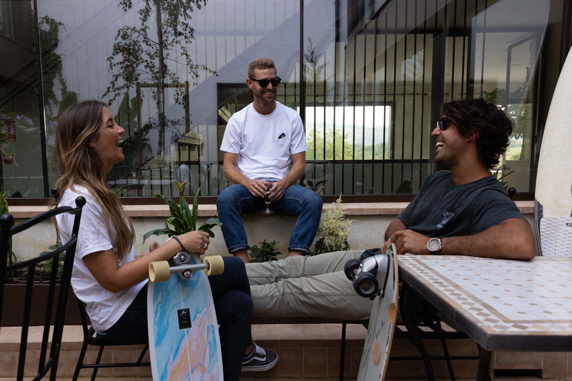 Tres personas charlando en una cafetería al aire libre con mesas, sillas y un edificio al fondo.