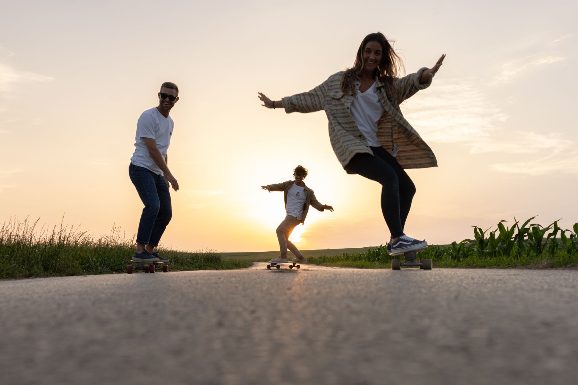 Tres personas patinan en monopatín por una carretera al atardecer; una de ellas está en primer plano, con campos de hierba al fondo.
