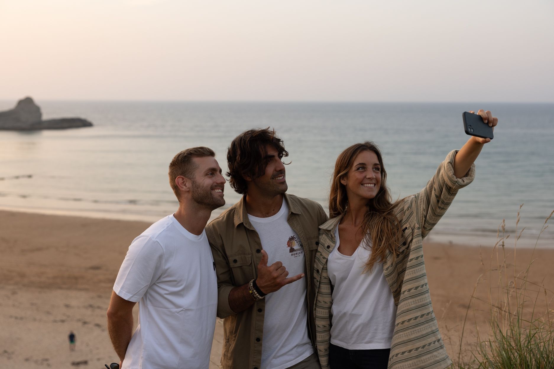 Tres amigas se toman una selfie en la playa al atardecer, sonriendo junto al océano.