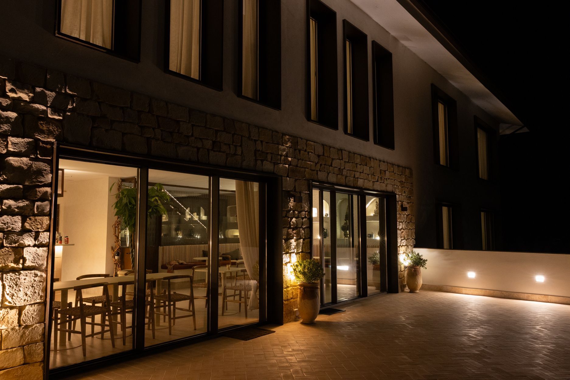 Vista nocturna de una moderna casa de piedra con puertas de cristal iluminadas y un patio cálidamente iluminado.