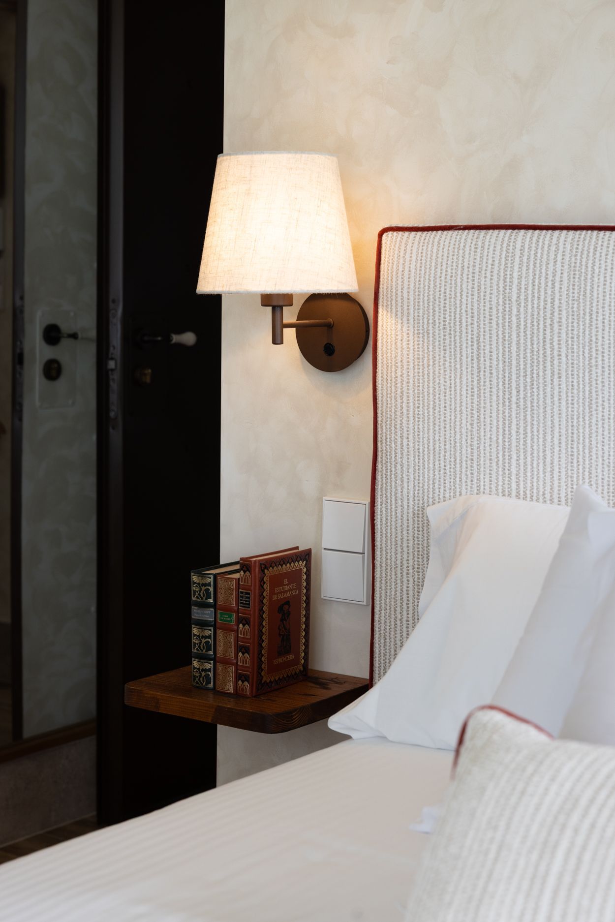 Dormitorio con cama blanca, cabecero texturizado, lámpara de pared y mesita de noche con libros.