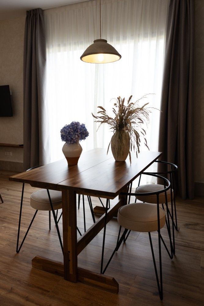 Mesa de comedor con dos sillas, jarrones con flores y una lámpara colgante junto a una ventana luminosa en una sala de estar.