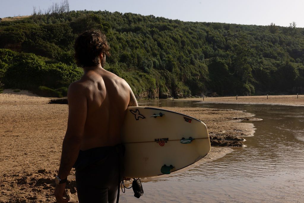 Un surfista sostiene una tabla de surf blanca en una playa de arena junto a aguas tranquilas y acantilados verdes.