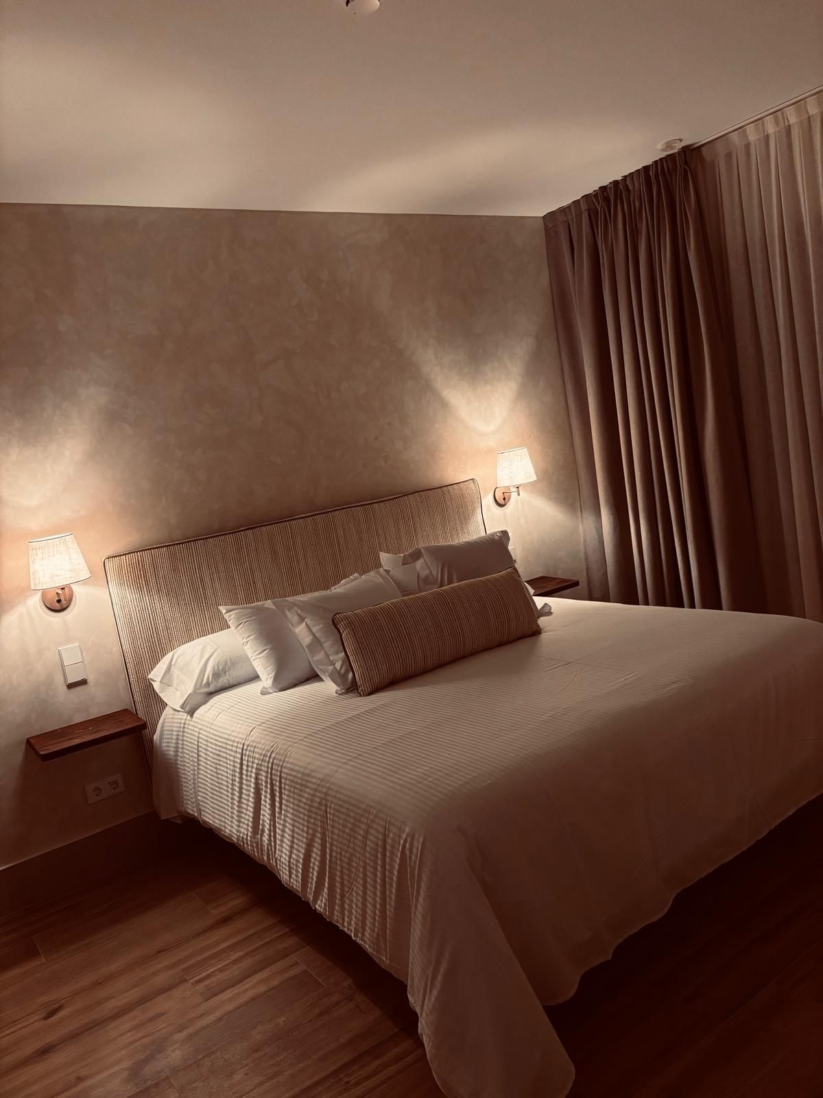 Habitación acogedora con cama, lámparas de pared y cortinas marrones en iluminación cálida.