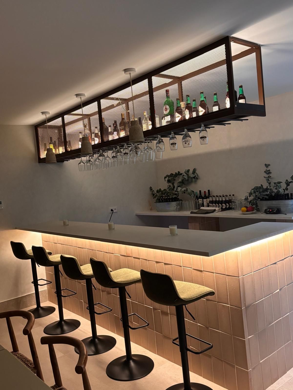 Elegante bar moderno con mostrador de azulejos blancos, taburetes negros y estante colgante para vasos bajo iluminación cálida.