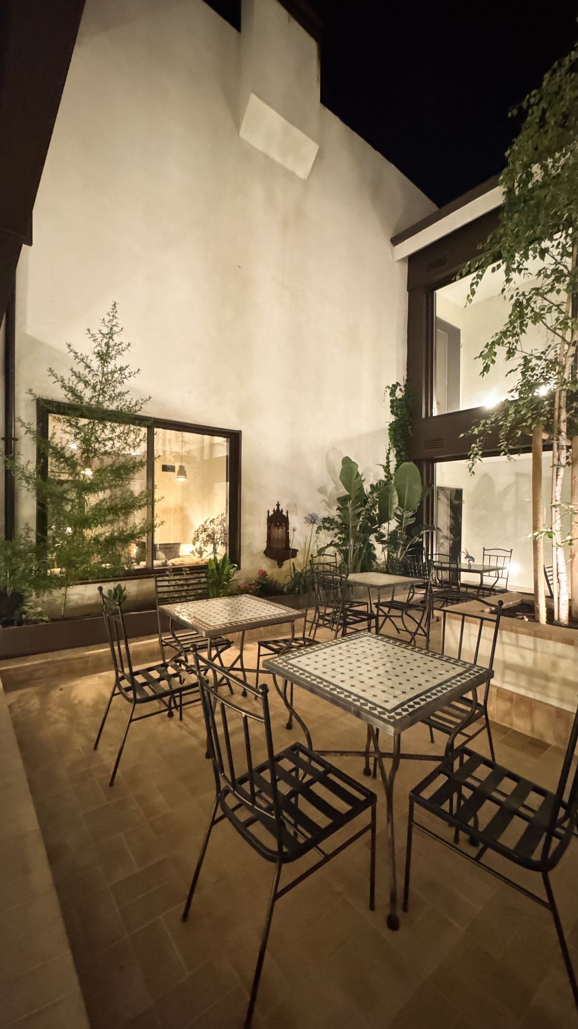 Comedor al aire libre con mesas y sillas de metal junto a un edificio moderno por la noche.