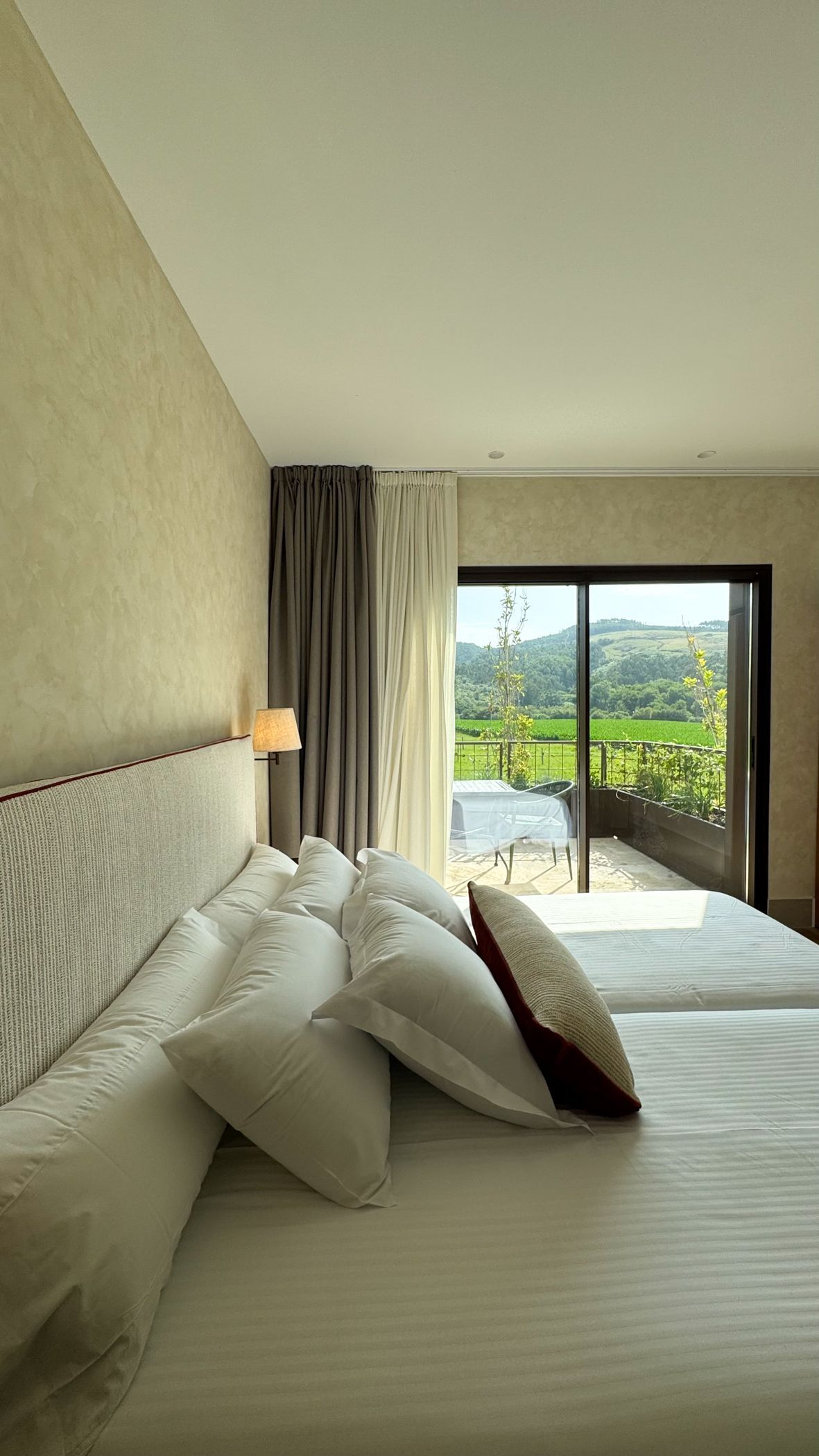 Habitación de hotel luminosa con cama blanca, cortinas beige y puerta corredera que da a un balcón con vistas a la vegetación.