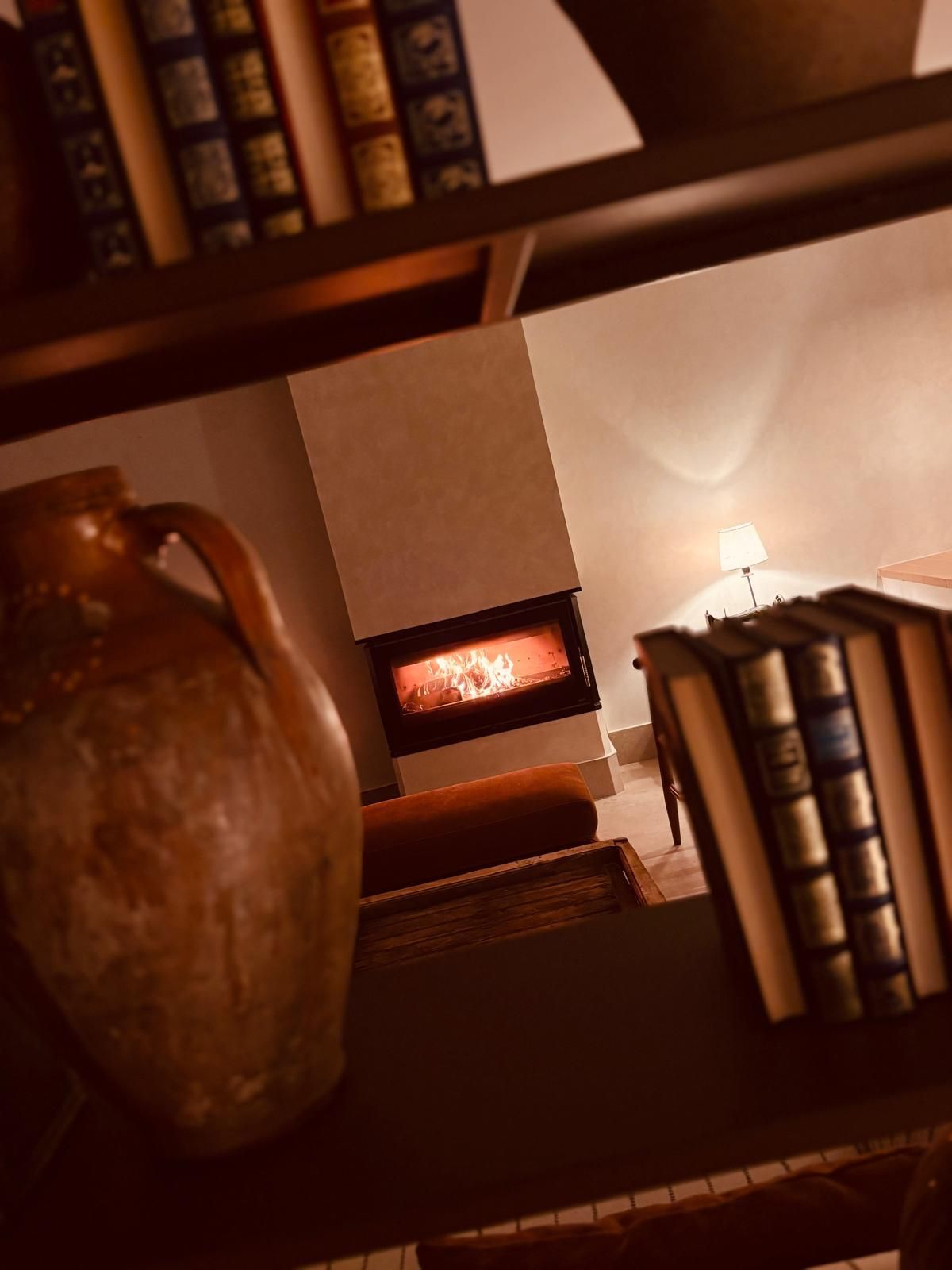 Acogedora biblioteca con estanterías, una chimenea crepitante y un gran jarrón de cerámica bajo una luz cálida.