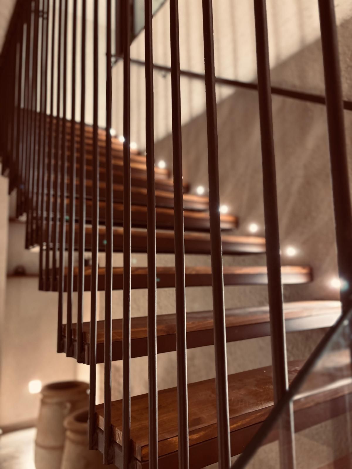 Escalera curva de madera con barandillas verticales y luces cálidas a lo largo de los escalones.