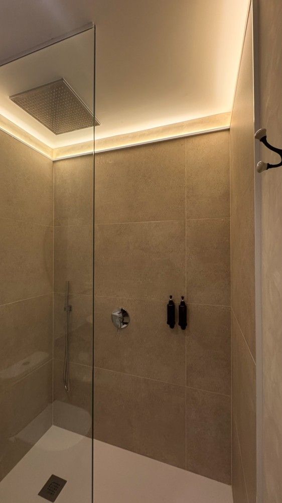 Ducha moderna con azulejos beige, pared de cristal, cabezal de ducha tipo lluvia e iluminación LED cálida en el techo.