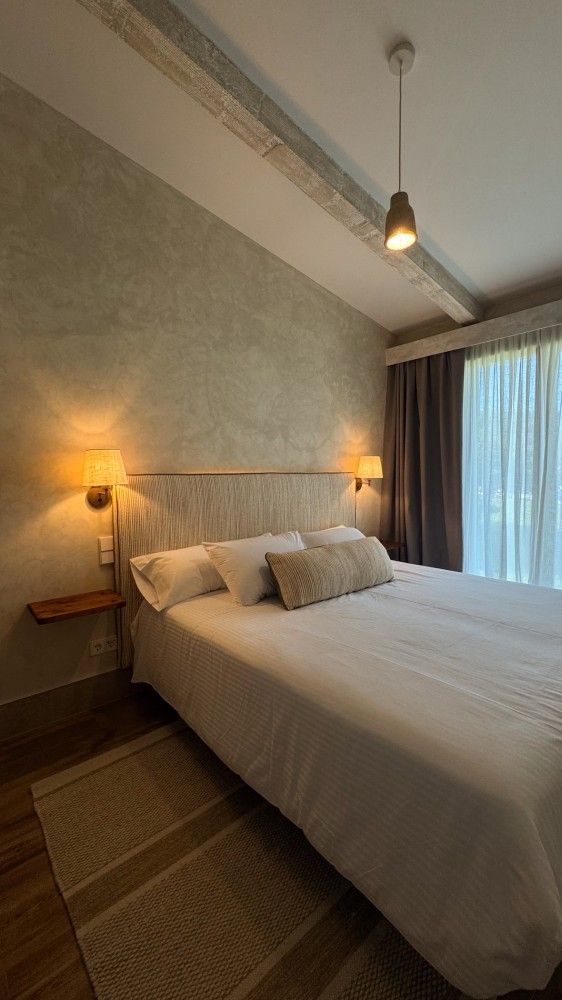 Habitación acogedora con una cama bien hecha, una lámpara de pared cálida, paredes con textura y cortinas que van del suelo al techo.