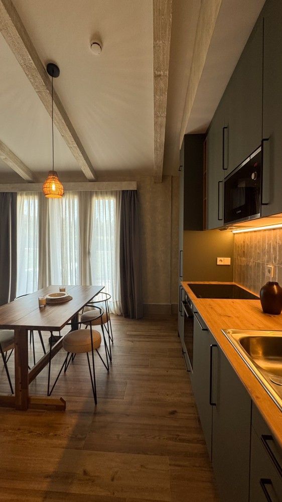 Cocina-comedor moderna con suelos de madera, iluminación naranja, mesa, sillas y cortinas junto a una ventana.