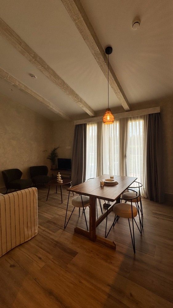 Comedor moderno con mesa de madera, sillas, lámpara colgante y cortinas transparentes junto a la ventana.