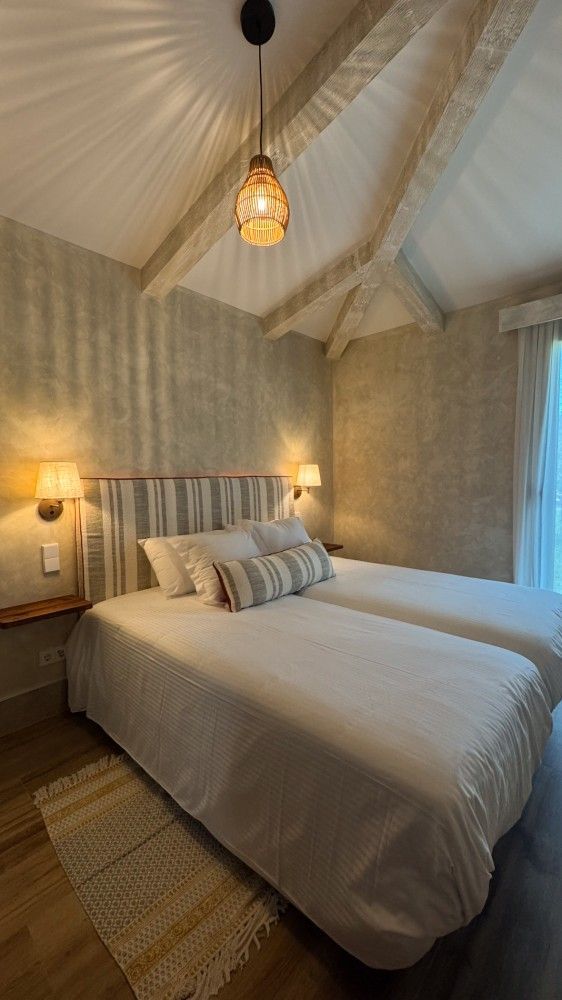 Acogedor dormitorio con cama blanca, lámpara colgante de mimbre y paredes neutras con textura.