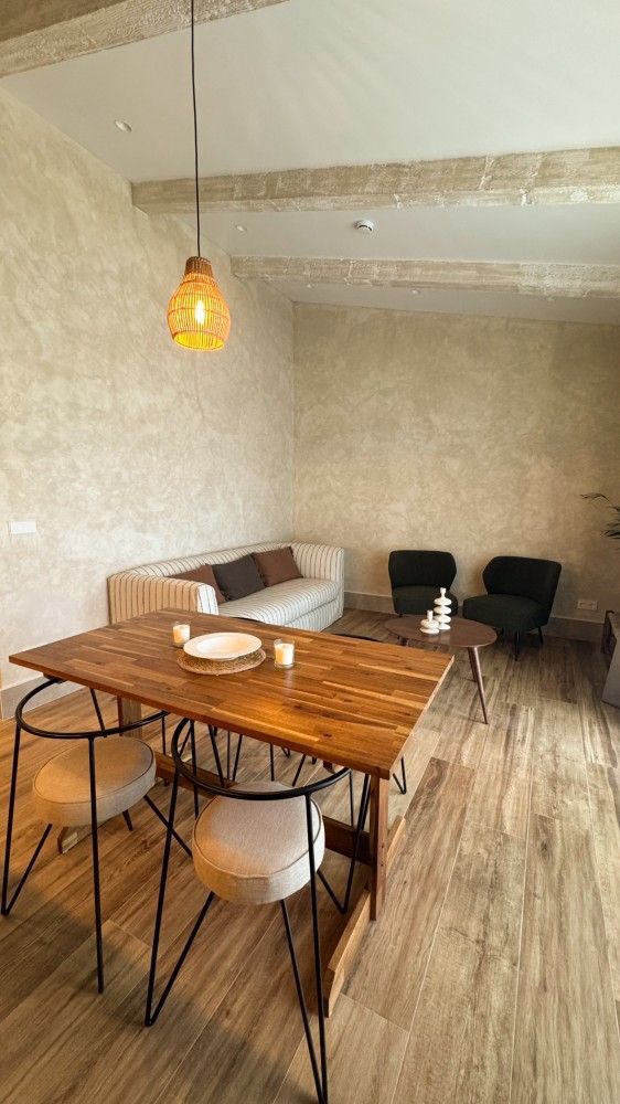 Acogedora sala de estar con mesa de madera, sofá beige, sillas negras, papel pintado estampado y lámpara colgante de color ámbar.