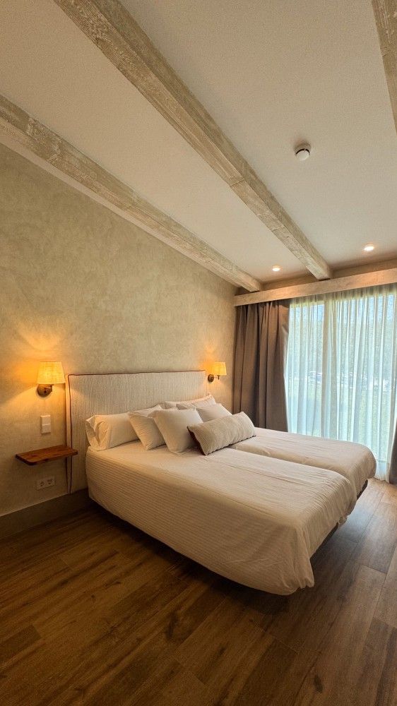Acogedora habitación con dos camas individuales, lámparas de pared cálidas, suelo de madera y cortinas transparentes junto a una gran ventana.