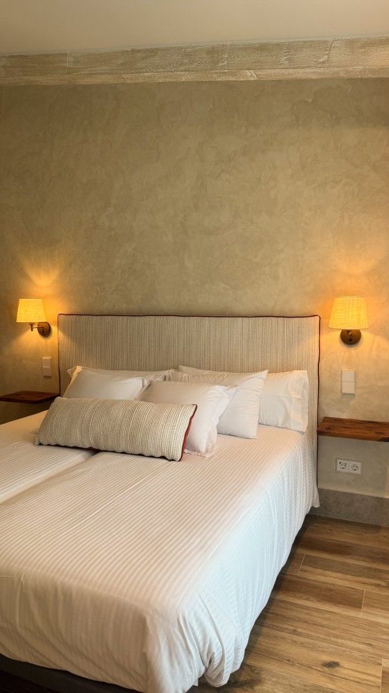 Cama bien hecha en un dormitorio con iluminación tenue, con dos lámparas de pared y un cabecero texturizado de color beige.