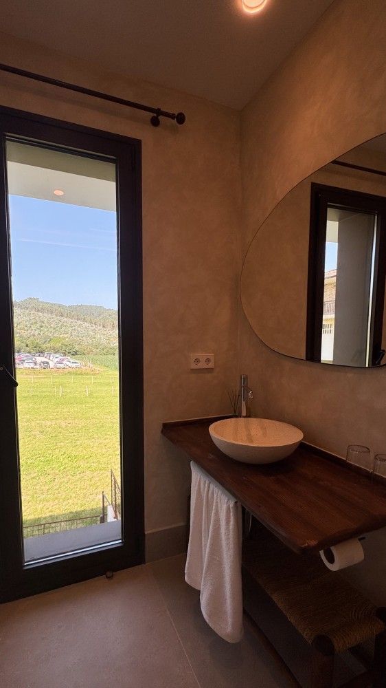 Baño minimalista con tocador de madera, lavabo redondo, espejo y puerta abierta a un paisaje verde exterior.