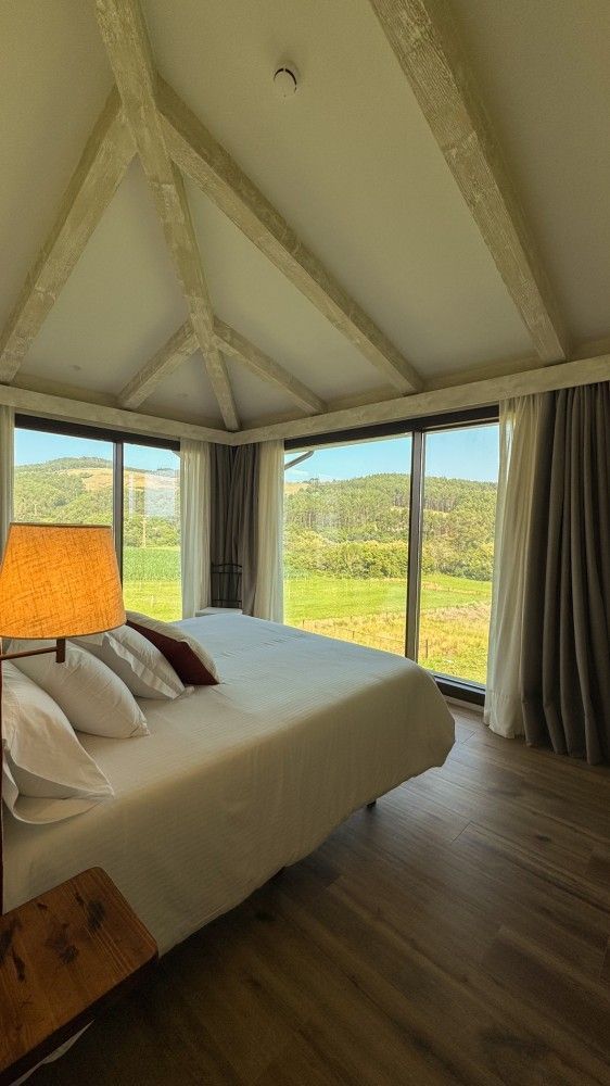 Habitación acogedora con cama, lámpara, suelo de madera y grandes ventanales con vistas a un paisaje verde.