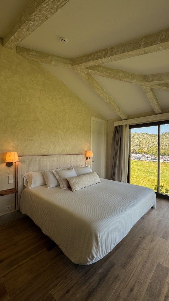 Habitación luminosa con cama blanca, lámparas cálidas, suelo de madera y gran ventanal con vistas a campos verdes.