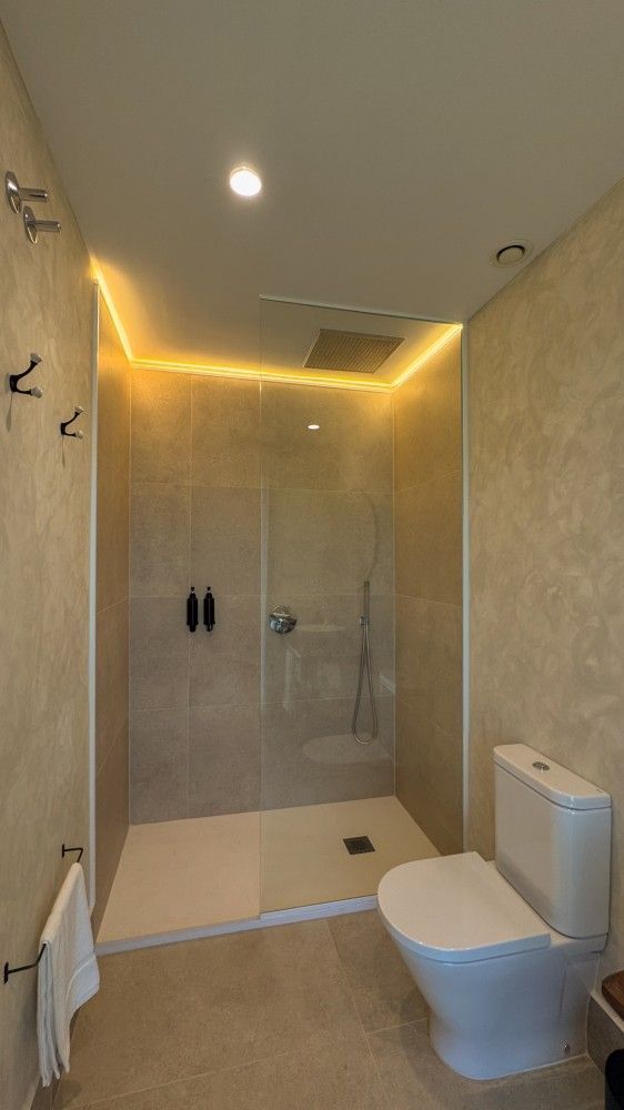 Baño moderno de color beige con ducha de cristal a ras de suelo, inodoro, toallero e iluminación cálida en el techo.