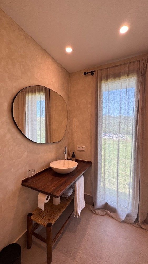 Pequeño tocador con espejo redondo, escritorio de madera, taburete blanco y ventana con cortinas transparentes con vegetación en el exterior.