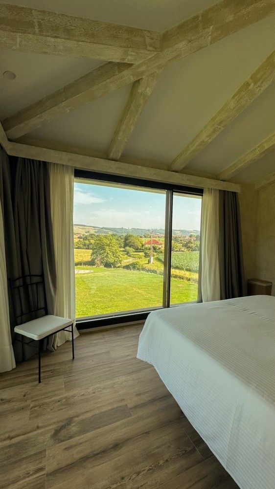 Habitación con un gran ventanal con vistas a campos verdes y colinas, suelo de madera y una cama blanca.