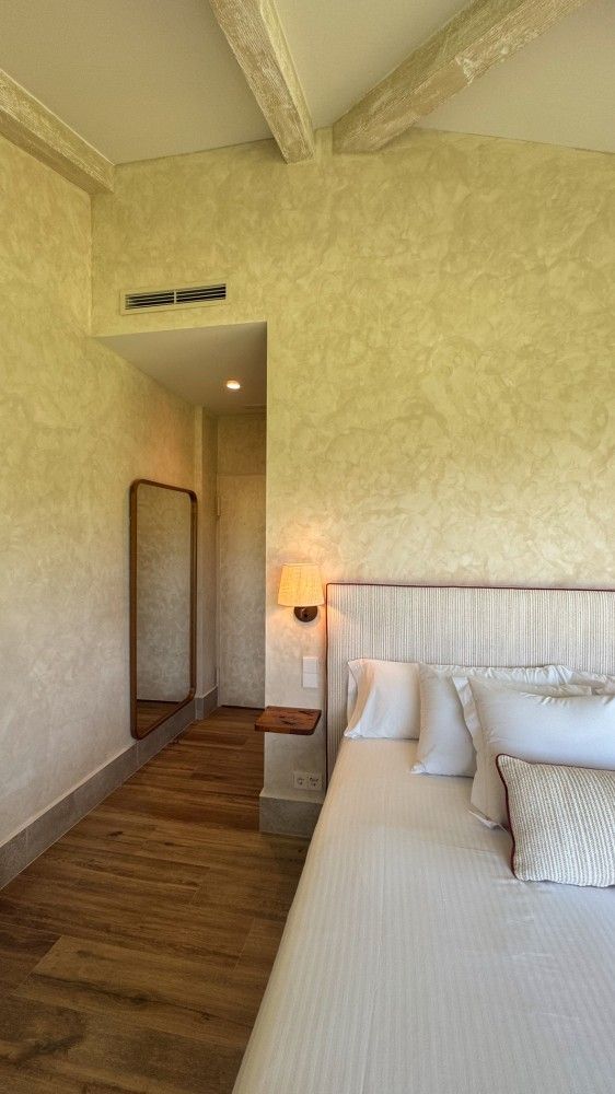 Habitación acogedora con cama blanca, suelo de madera, paredes texturizadas y lámpara de noche cálida.
