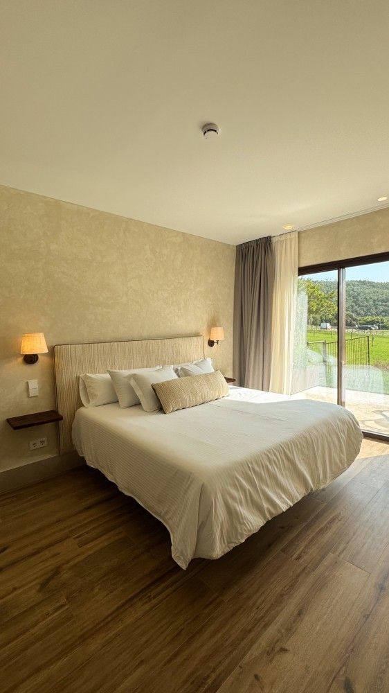 Habitación luminosa y moderna con cama, lámpara de noche, suelo de madera y puertas que dan a un balcón con vistas a la vegetación.
