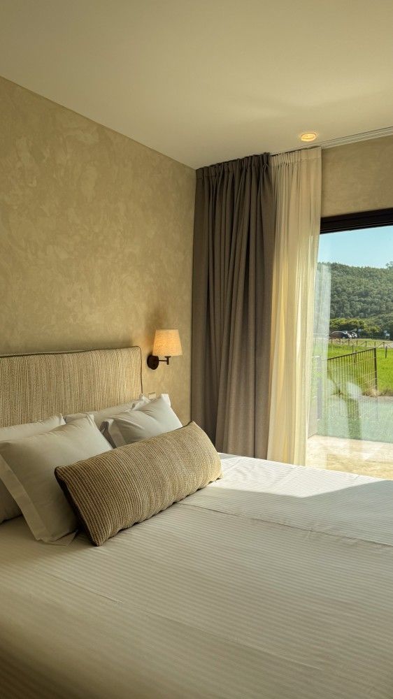 Elegante dormitorio con una cama impecablemente hecha, paredes de color beige y vistas a la vegetación desde la ventana.