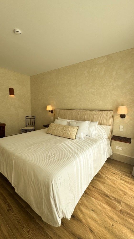 Cama bien hecha en una habitación de hotel de color beige con lámparas de noche y suelo de madera.