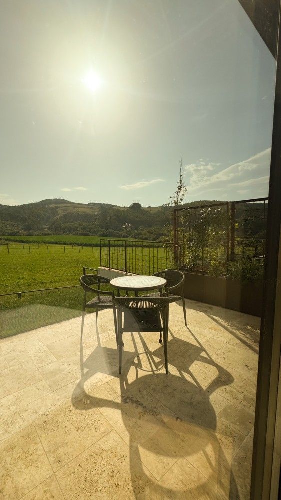 Patio soleado con mesa redonda y sillas junto a un campo verde y colinas a lo lejos.