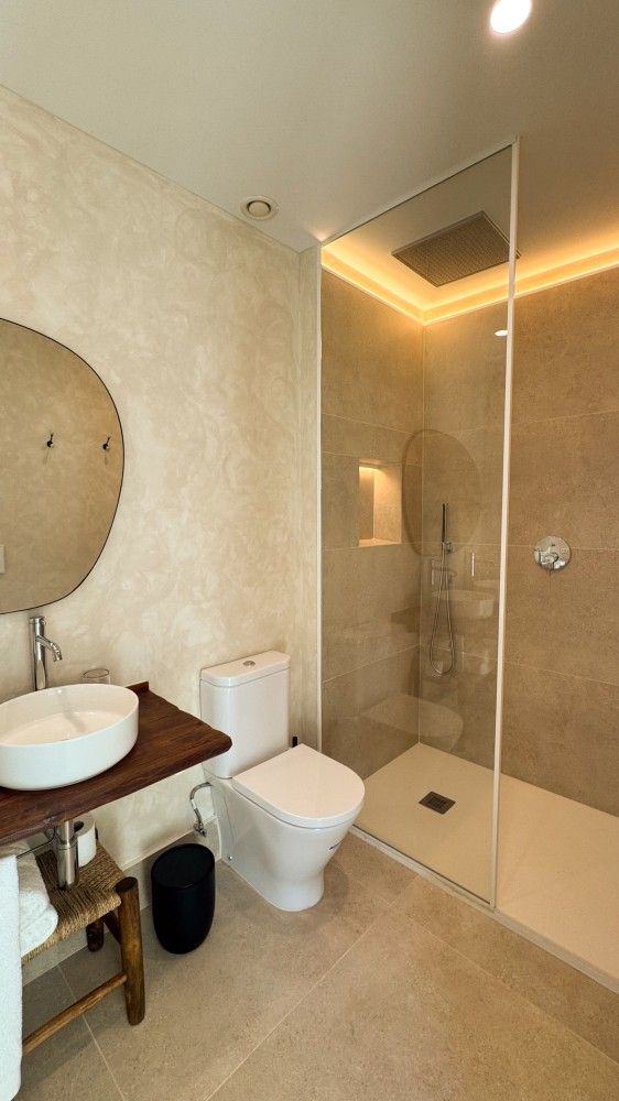 Baño moderno de color beige con lavabo, inodoro y cabina de ducha de cristal con iluminación cálida.
