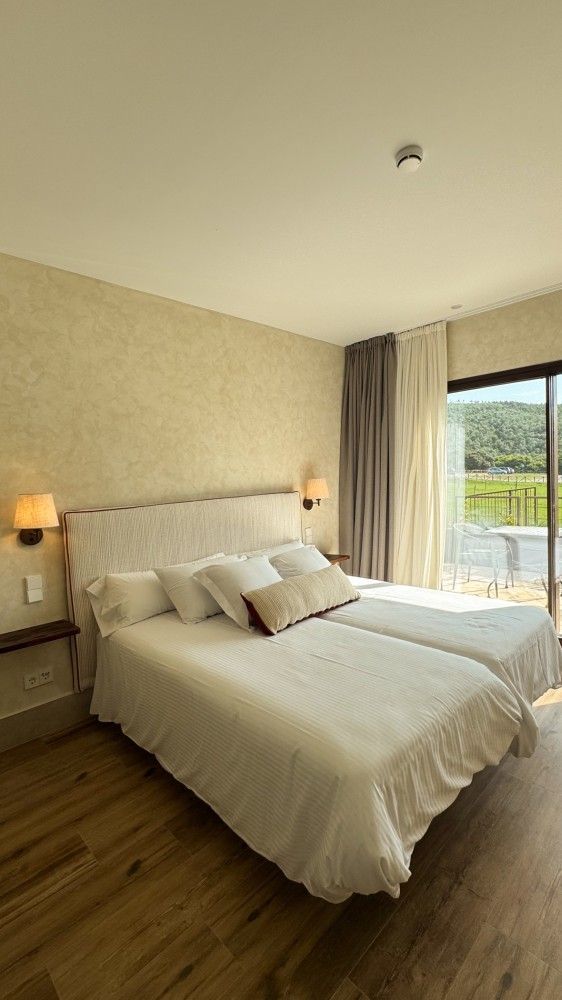 Habitación luminosa con cama blanca bien hecha, lámparas de noche, suelo de madera y vistas al balcón a través de puertas correderas.