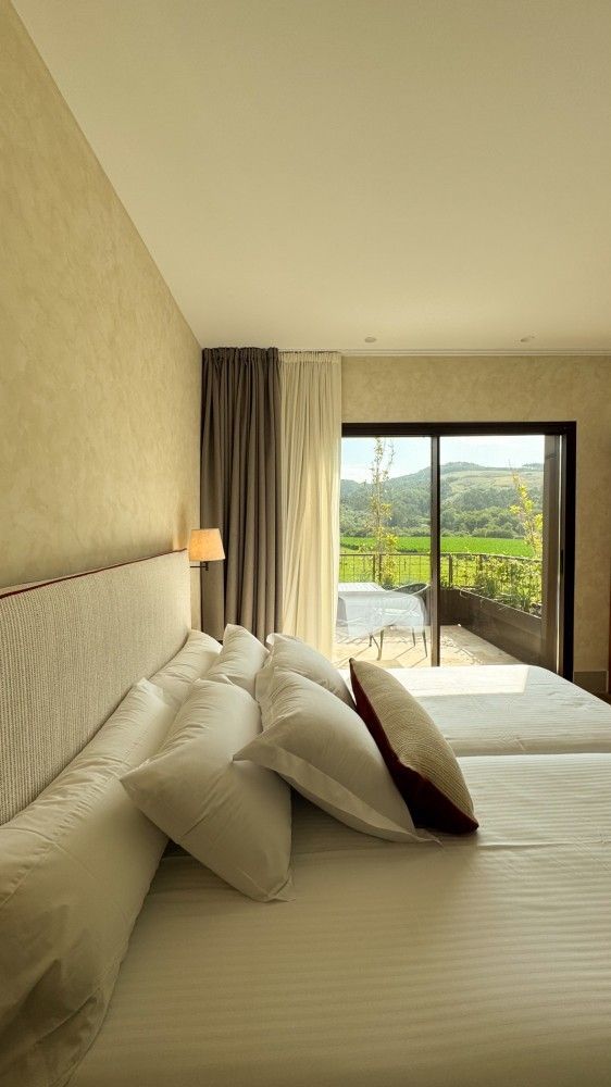 Habitación de hotel con cama impecablemente hecha, paredes de color beige y puertas corredizas de cristal que dan a un paisaje verde.