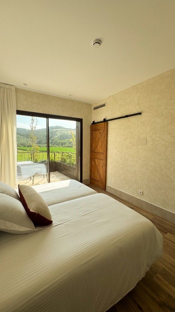 Habitación luminosa con dos camas blancas, puertas correderas de cristal y vistas a las verdes colinas del exterior.