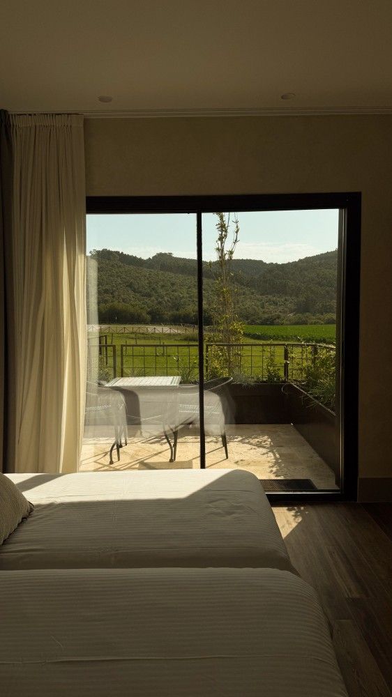 Habitación con cama frente a una puerta corrediza de cristal que da a un patio y a las verdes colinas al atardecer.